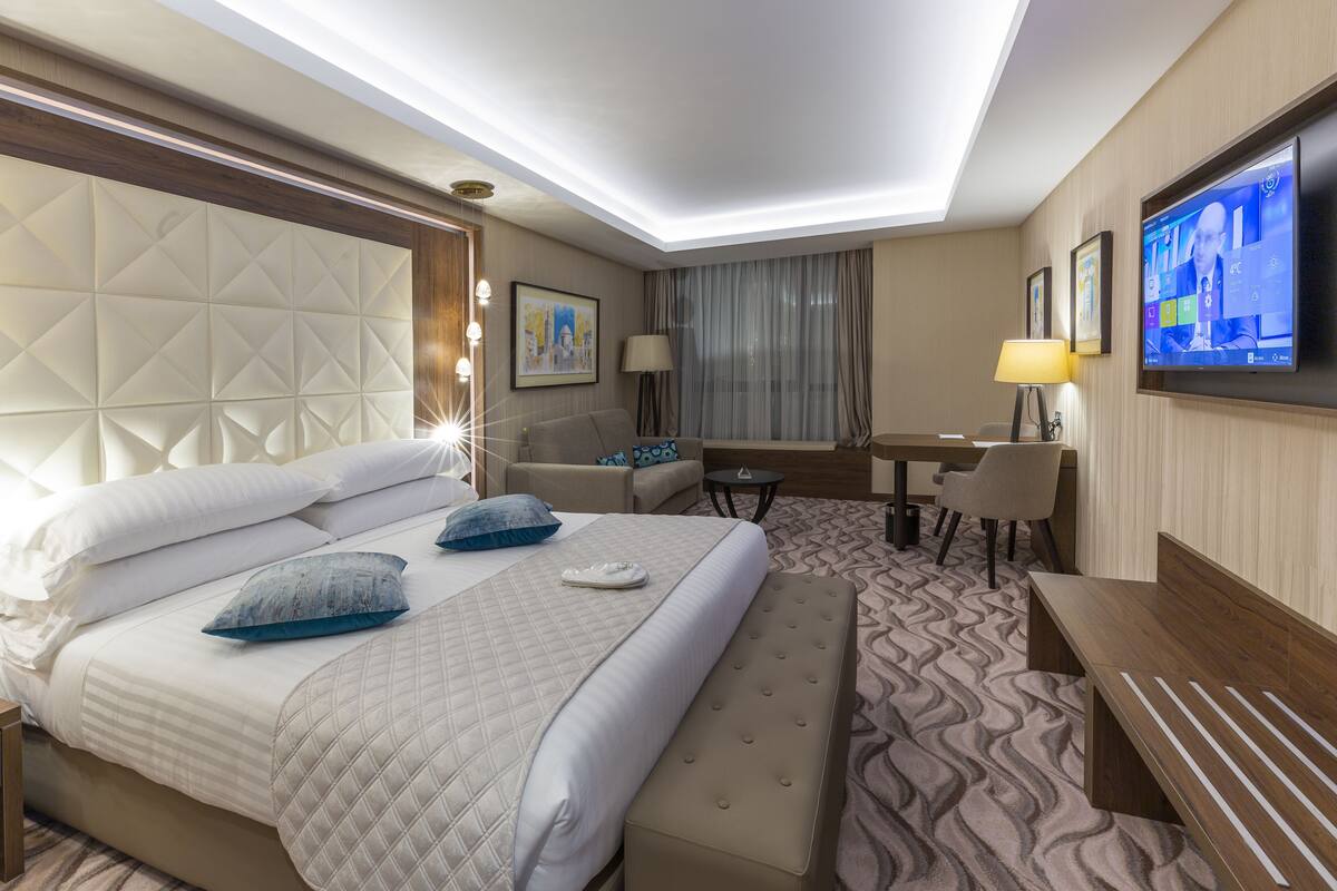 A hotel room with a large bed, a desk, a chair, a TV, and a patterned carpet.