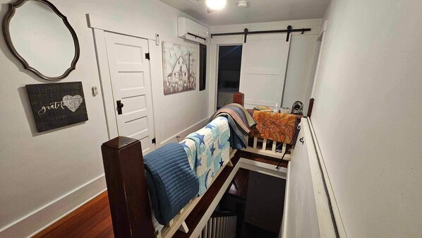A narrow hallway with a wooden staircase, a bed with a blue quilt, a wall-mounted mirror, and a door with a small picture. - Relaxing country setting perfect for that special get away!! (Beatrice)