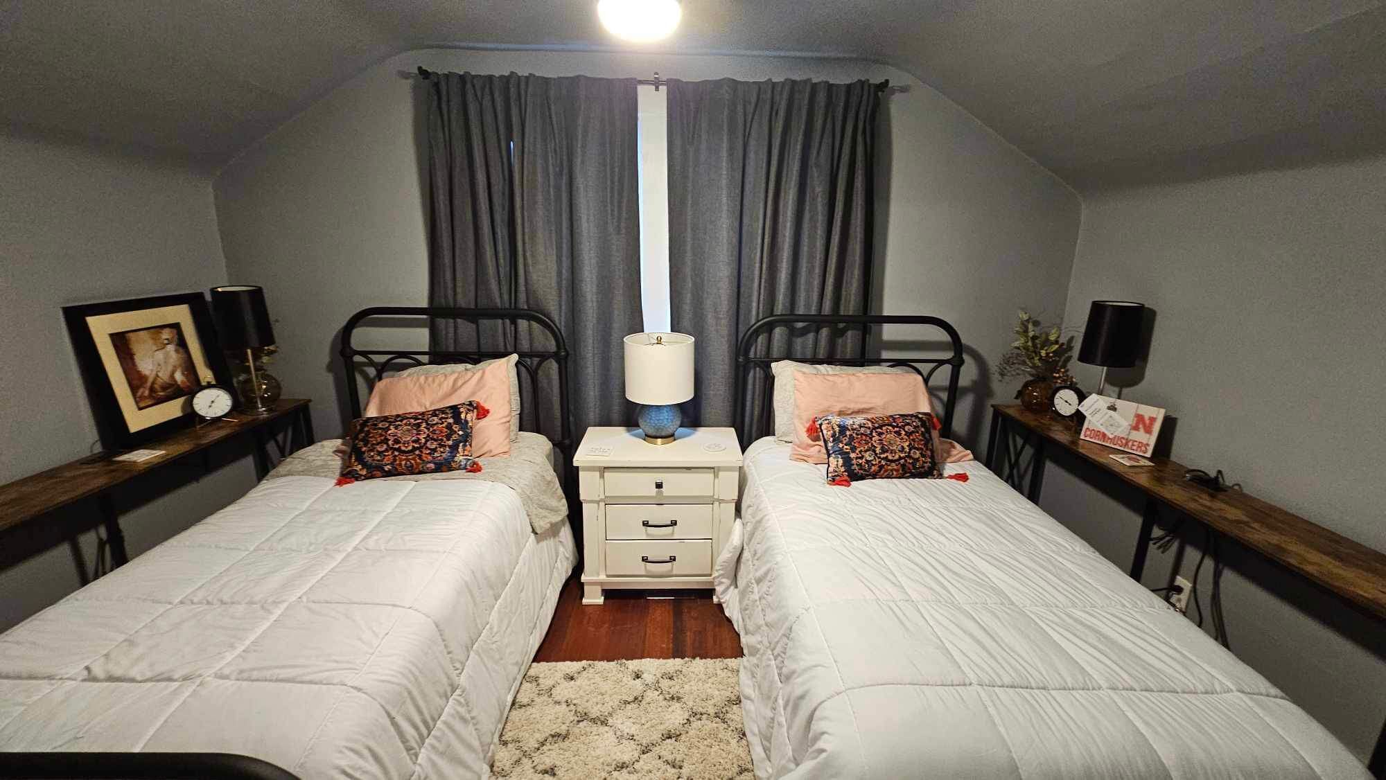 Une chambre à deux lits simples, avec deux tables de chevet équipées de lampes, une table de nuit, un tableau encadré et une horloge.