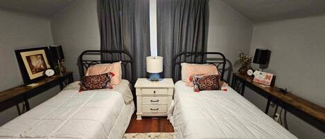 Une chambre à deux lits simples, avec deux tables de chevet équipées de lampes, une table de nuit, un tableau encadré et une horloge.