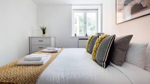 A modern bedroom with a large bed, a bedside table, a window, and a framed picture on the wall. - The Waterloo Flats (London)