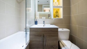 A modern bathroom with a glass shower enclosure, a white bathtub, a wooden vanity with a sink, and a toilet. - The Waterloo Flats (London)