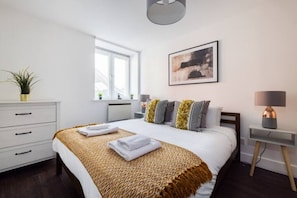 A modern hotel room with a large bed, a bedside table with a lamp, a window, and a wall-mounted picture.