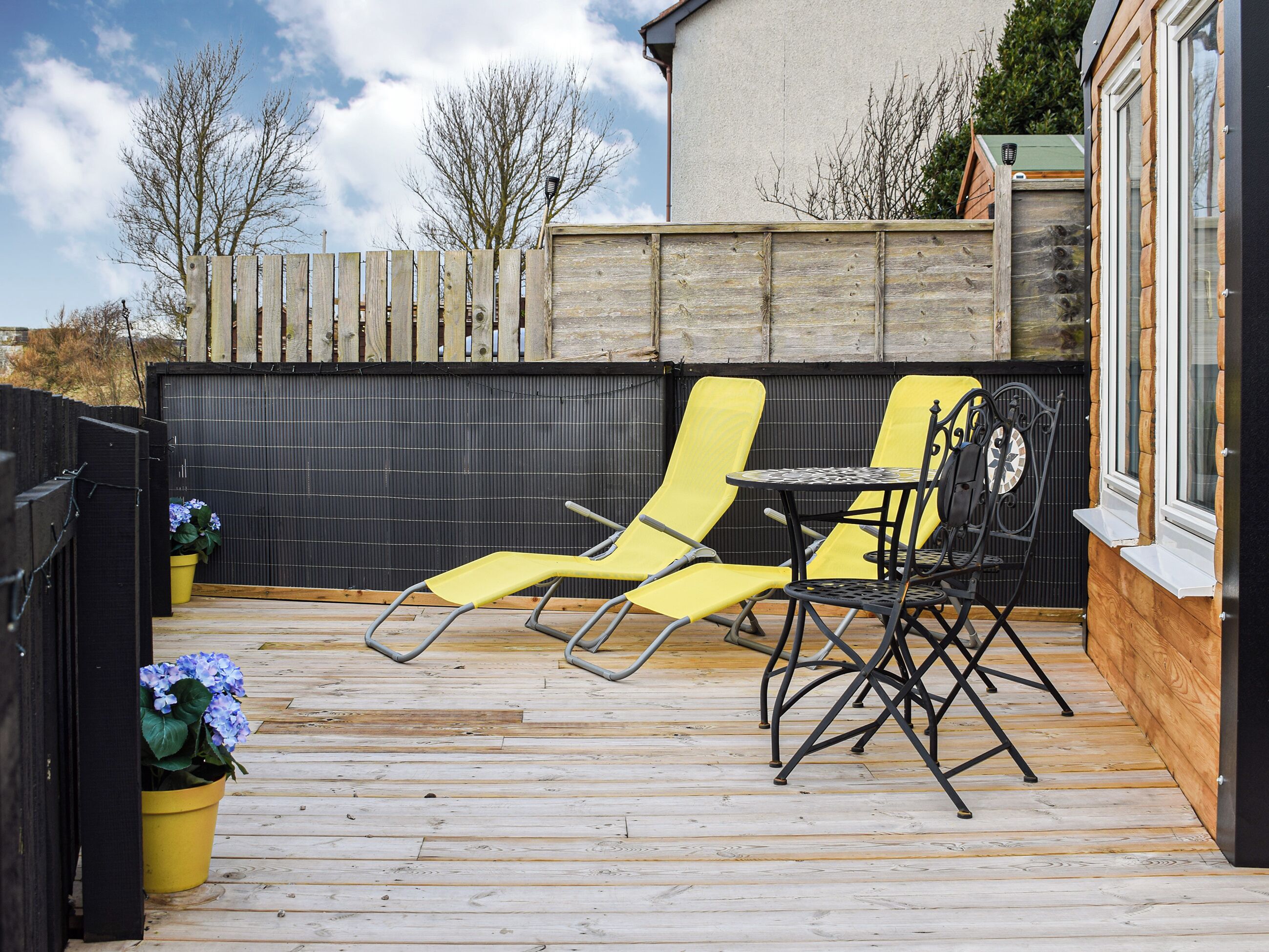 Eine Holzterrasse mit gelben Stühlen und einem Tisch, ein schwarzer Zaun und ein Gebäude im Hintergrund.