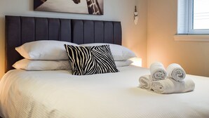 Ein ordentlich bezogenes Bett mit weißen und gestreiften Kissen, einem Kissen mit ZEBRA-Muster und aufgerollten Handtüchern. Über dem Bett hängt ein Gemälde mit einem Giraffenmotiv.