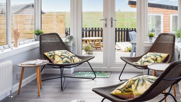 Ein modernes Zimmer mit Korbstühlen und einem kleinen Tisch, große Fenster mit Blick auf eine Holzterrasse und einen Zaun.