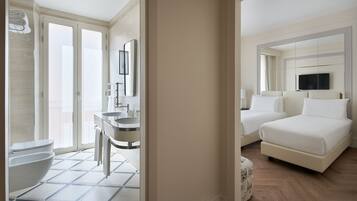 A hotel room with a bathroom featuring a toilet, bidet, and sink. There is a bed with two pillows and a television mounted above it.