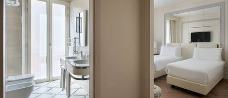 A hotel room with a bathroom featuring a toilet, bidet, and sink. There is a bed with two pillows and a television mounted above it.