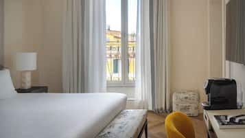 A hotel room with a bed, a bedside lamp, a window with curtains, a small table with a coffee maker, and a suitcase.