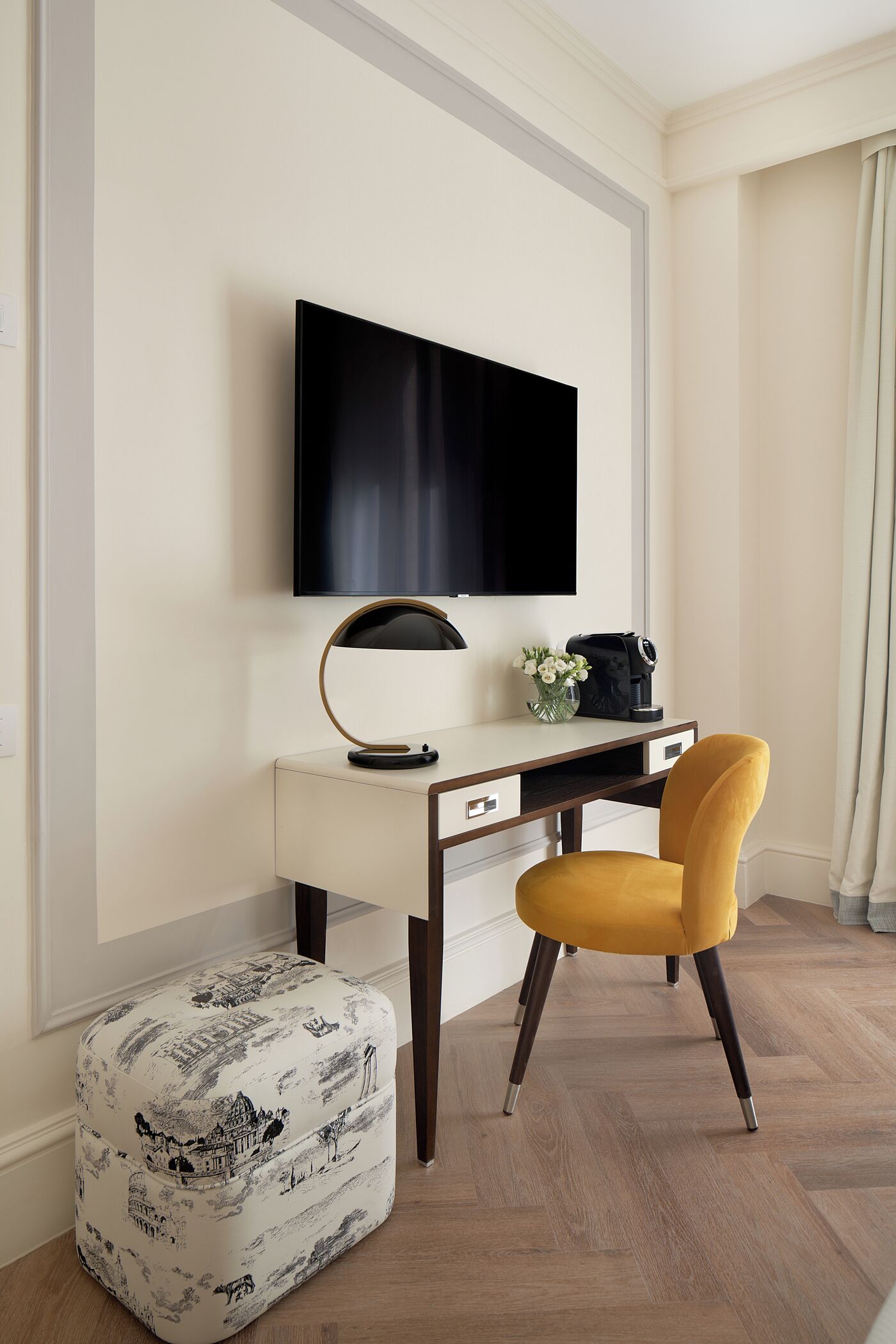 A modern hotel room with a flat-screen TV mounted on the wall, a desk with a lamp, a coffee maker, and a yellow chair. There is a decorative ottoman with a black and white design.