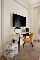 Een moderne hotelkamer met een flatscreen televisie aan de muur, een bureau met een lamp, een koffiezetapparaat en een gele stoel. Er staat een decoratieve poef met een zwart-wit ontwerp.