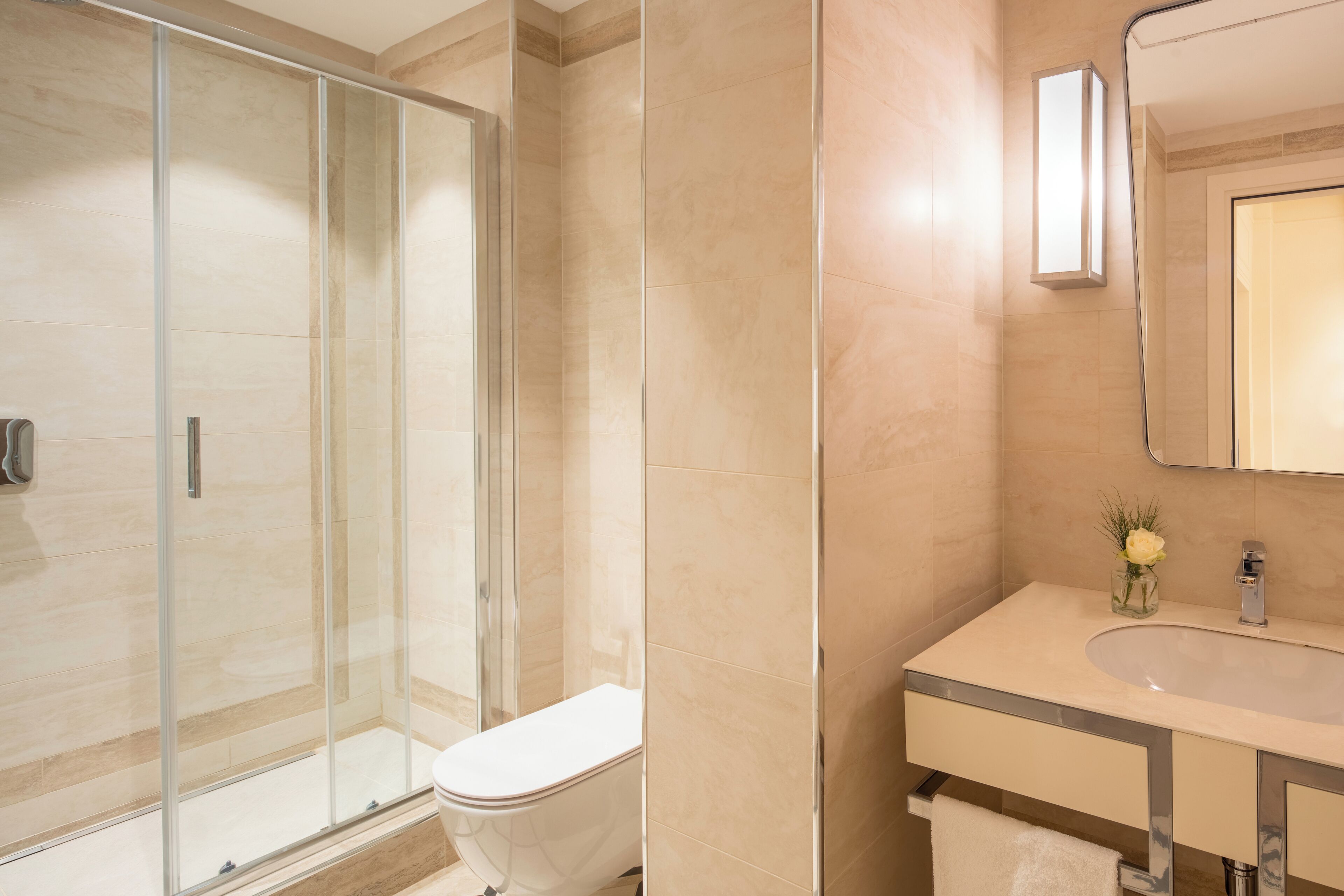 A modern bathroom with a glass-enclosed shower, a white toilet, and a vanity with a mirror and a small vase of flowers.