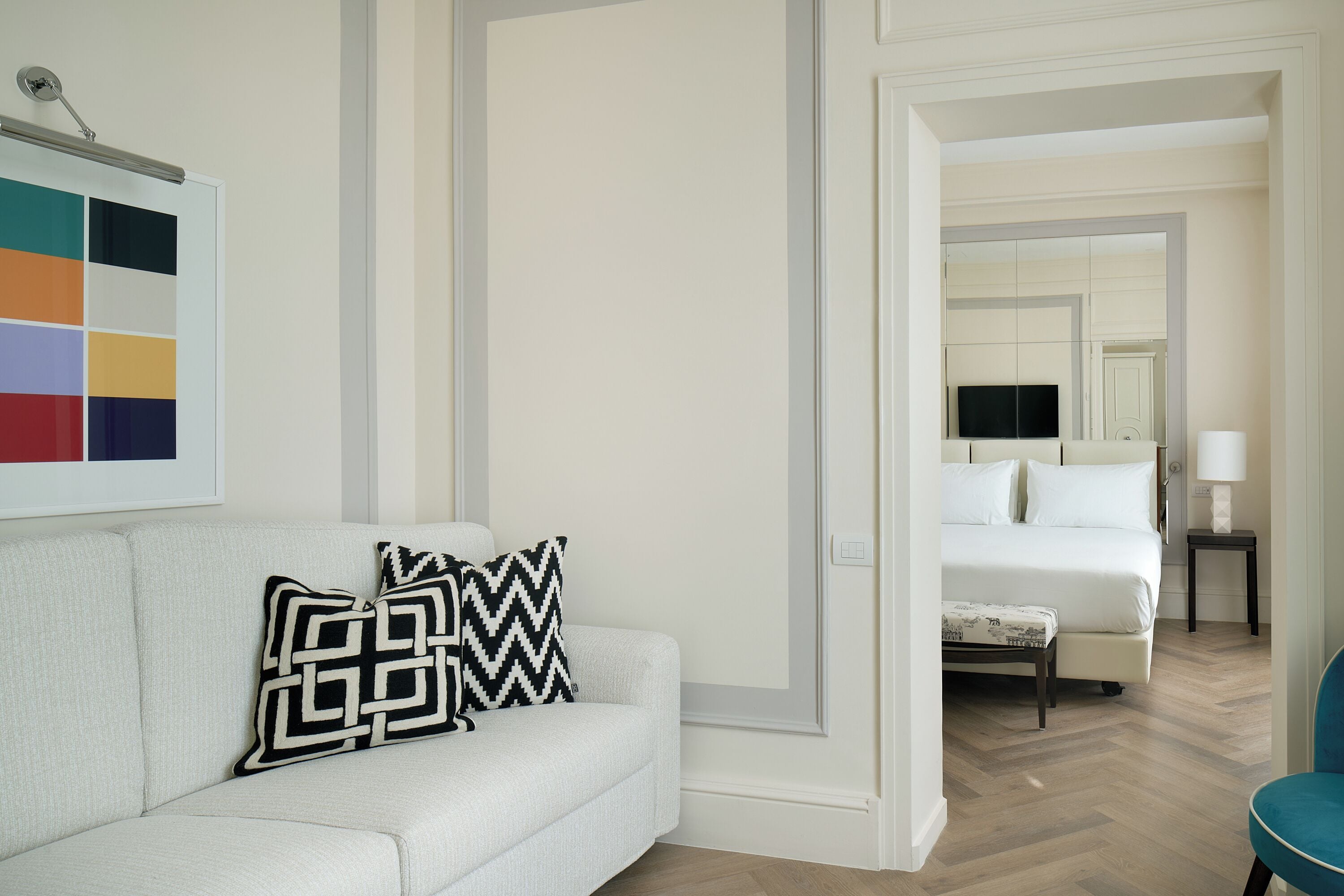 Una camera d'albergo con un divano bianco, un cuscino decorato, un letto con biancheria bianca, una televisione su un mobile e un comodino con una lampada.