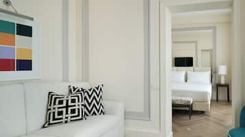 Una camera d'albergo con un divano bianco, un cuscino decorato, un letto con biancheria bianca, una televisione su un mobile e un comodino con una lampada.