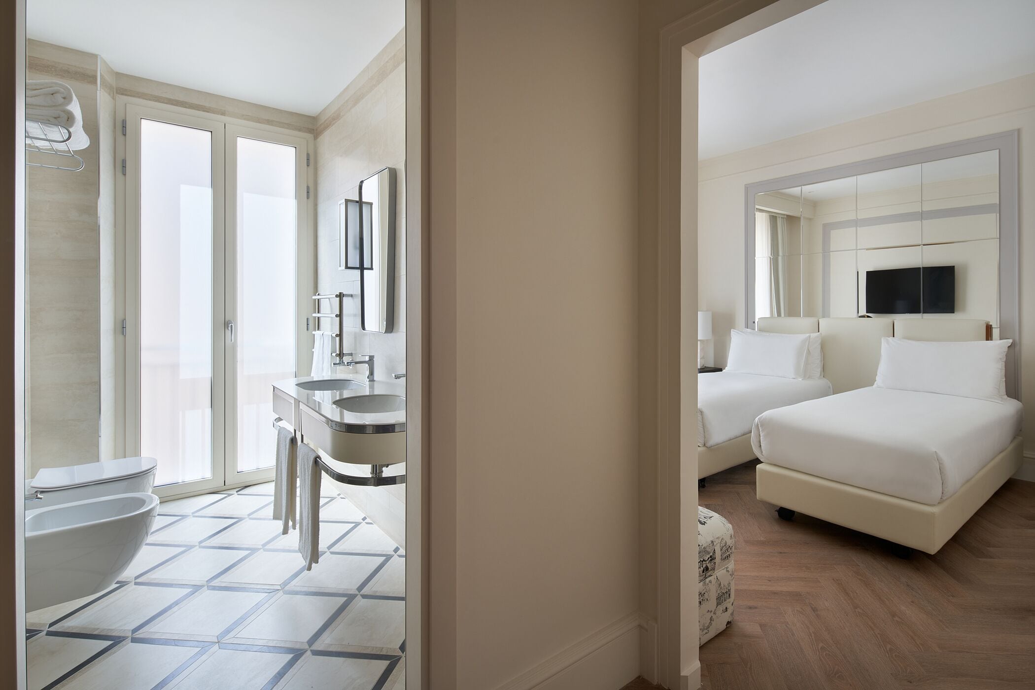 Camera d'albergo moderna con bagno dotato di WC, bidet e lavandino. Presenta un letto con due cuscini e una televisione montata sopra.