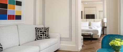 A modern hotel room with a white sofa, a blue armchair, a bed with white linens, and a large mirror reflecting the room.