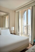 Quarto de hotel com uma cama grande, mesa de cabeceira com abajur, janela com cortinas e varanda com guarda-corpo.