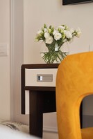 Una habitación con una mesita de noche de madera, un florero con flores blancas y una silla amarilla.