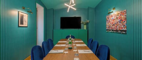 A conference room with a long table, blue chairs, a projector screen, and a large poster on the wall.