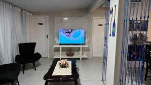 Ein Wohnzimmer mit Fernseher, zwei schwarzen Sesseln, einem Couchtisch mit Blumen und einem blauen Zaun.
