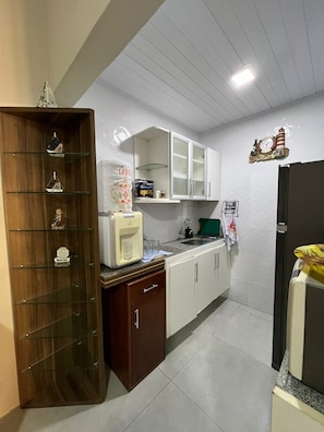 Une cuisine compacte avec des meubles blancs, une étagère en bois ornée d’objets décoratifs, une machine à café et une horloge au mur.