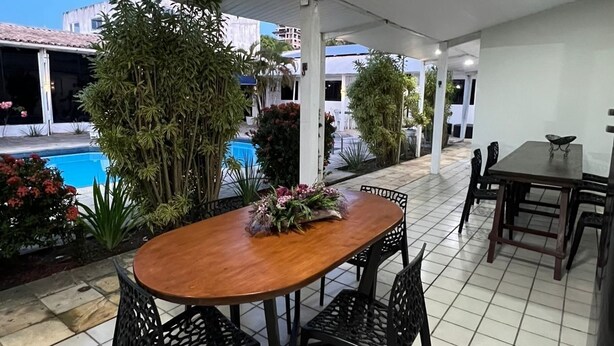Un patio al aire libre con mesa y sillas de madera, rodeado de plantas en macetas y una piscina.