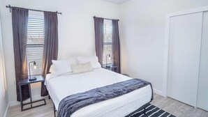 Ein Schlafzimmer mit einem Bett, zwei Nachttischen, einem Hängespiegel und einem Fenster mit Vorhängen.