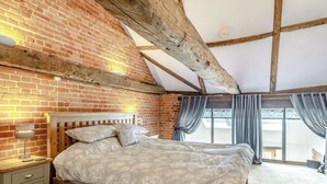Une chambre avec un mur en briques, des poutres en bois au plafond, un lit avec une literie à motifs floraux, une table de chevet avec une lampe, et une vue dégagée grâce à de grandes fenêtres.