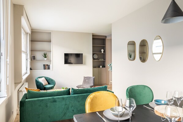 A modern living room with a green sofa, yellow chair, and a dining table set with wine glasses and plates.