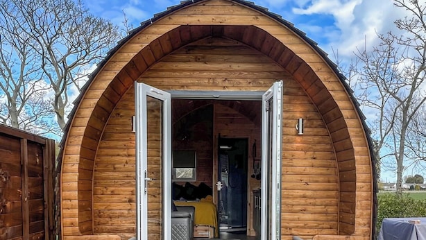 A wooden cabin with an arched entrance, a small porch, and a visible interior with a bed and a table.