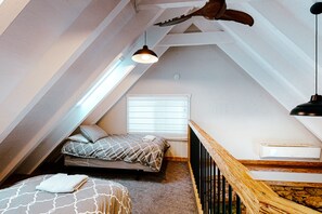 Une chambre mansardée chaleureuse avec un plafond en pente, un lit avec une literie à motifs, une fenêtre avec des stores et une rambarde en bois.