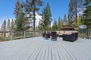 Une terrasse en bois, agréée de mobilier d’extérieur, entourée de grands arbres et bordée d’un bâtiment équipé d’une antenne parabolique.