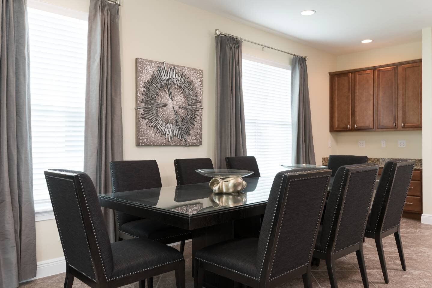 Un comedor con mesa de vidrio y seis sillas tapizadas, una pieza decorativa de metal en la pared, y una zona de cocina con armarios de madera.
