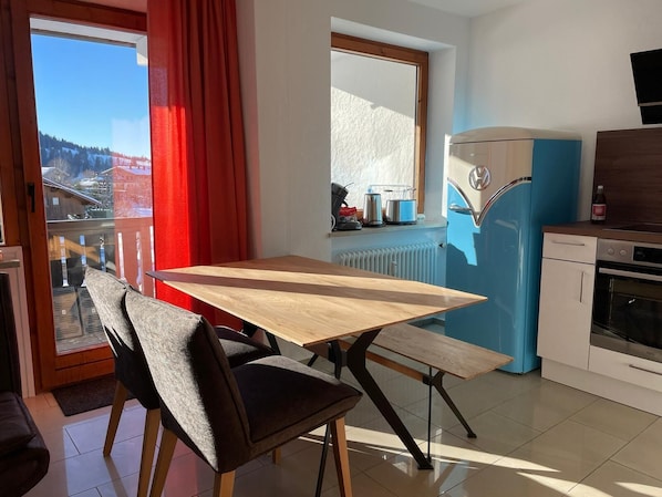 Une cuisine moderne avec un réfrigérateur bleu, une table à manger en bois avec des chaises, et une fenêtre donnant sur un paysage enneigé.