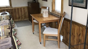 Une pièce chaleureuse avec une table à manger en bois, deux chaises, un banc recouvert de couvertures, un tableau encadré au mur et une fenêtre donnant sur un beau paysage.