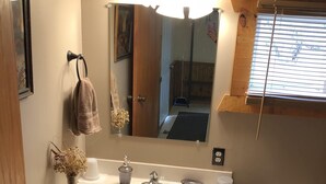 Une salle de bain avec un meuble-lavabo en bois, un lavabo blanc, un miroir, un porte-serviettes, des toilettes et une fenêtre avec des stores.