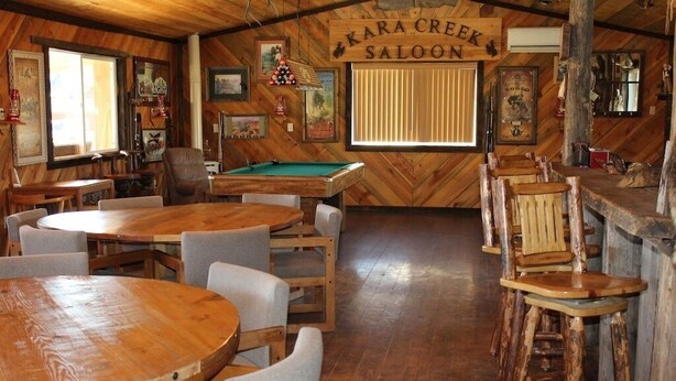 Un coin bar rustique avec des tables et des chaises en bois, une table de billard et une enseigne indiquant « KARA CREEK SALOON ».