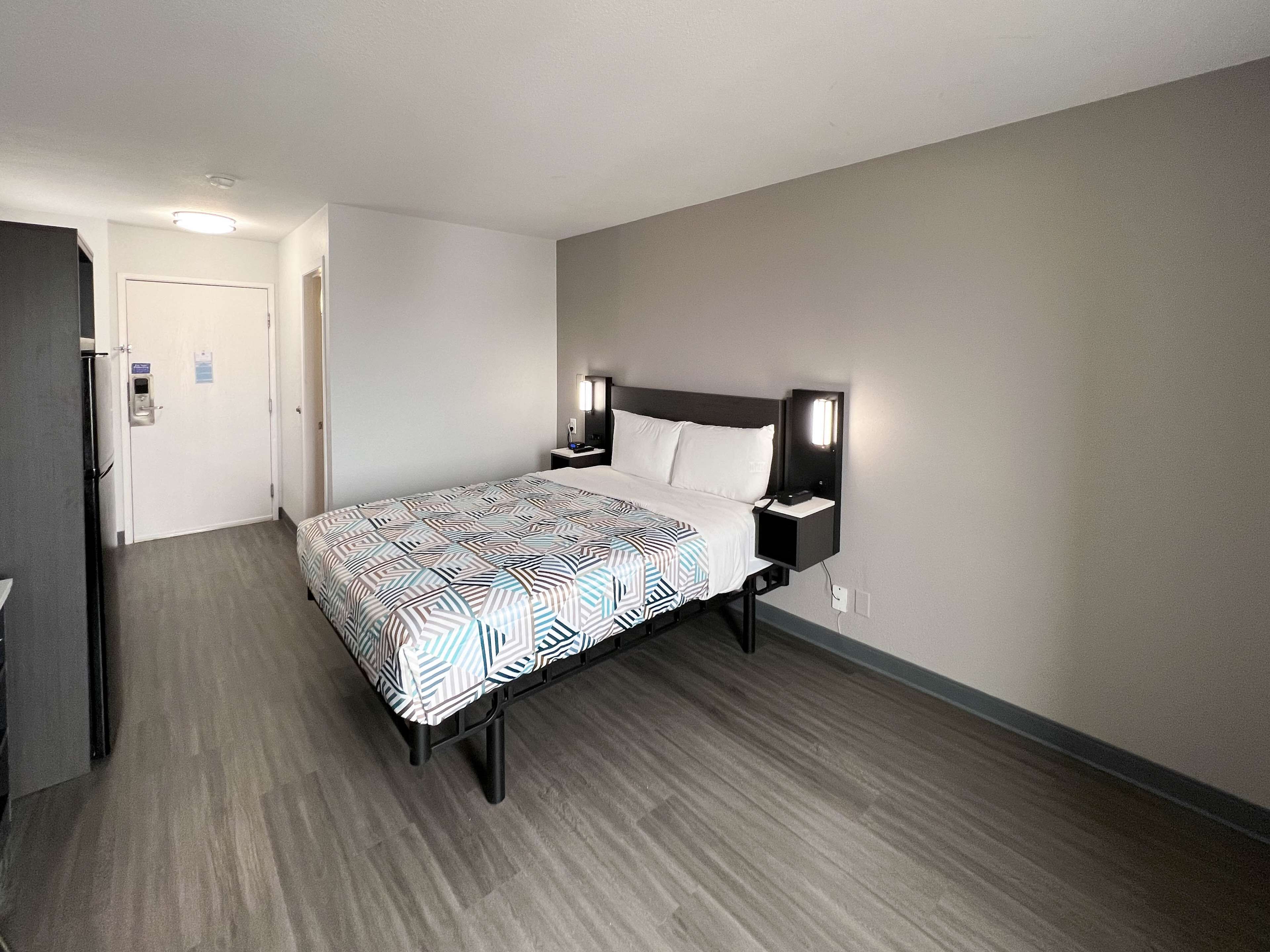 A hotel room with a bed, bedside tables, a nightstand, a wall-mounted lamp, and a door.
