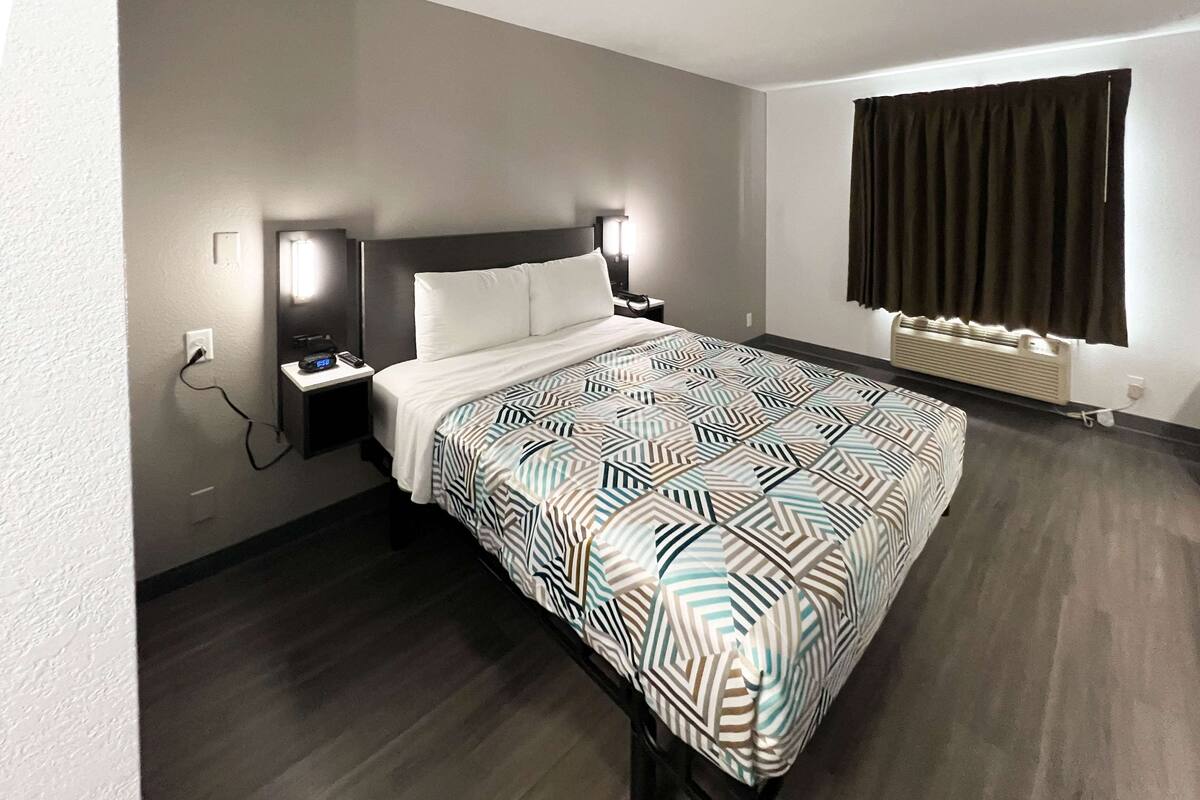 A hotel room with a large bed, two bedside tables with lamps, a window with curtains, and a wall-mounted heater.