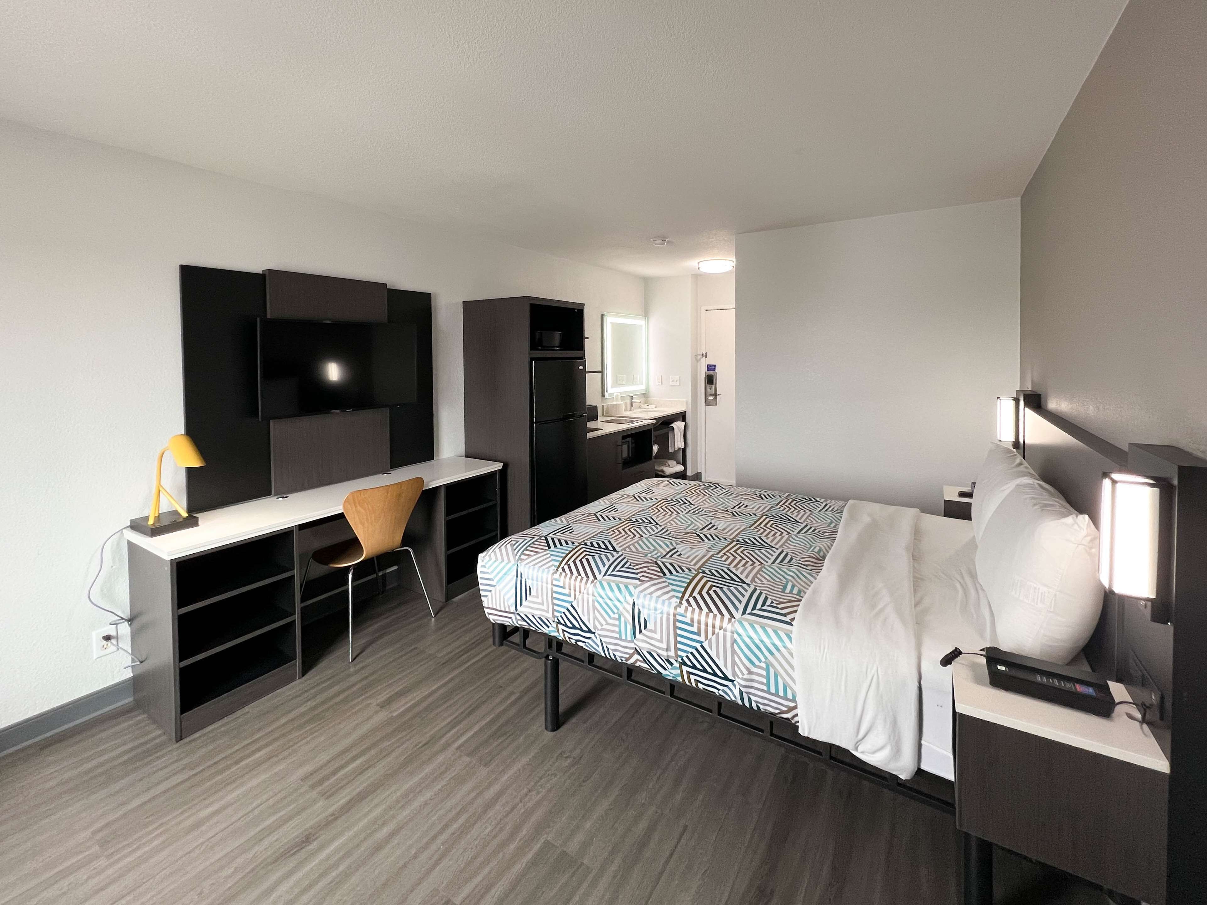 A modern hotel room with a bed, a desk with a chair, a television, a mirror, and a small kitchenette.