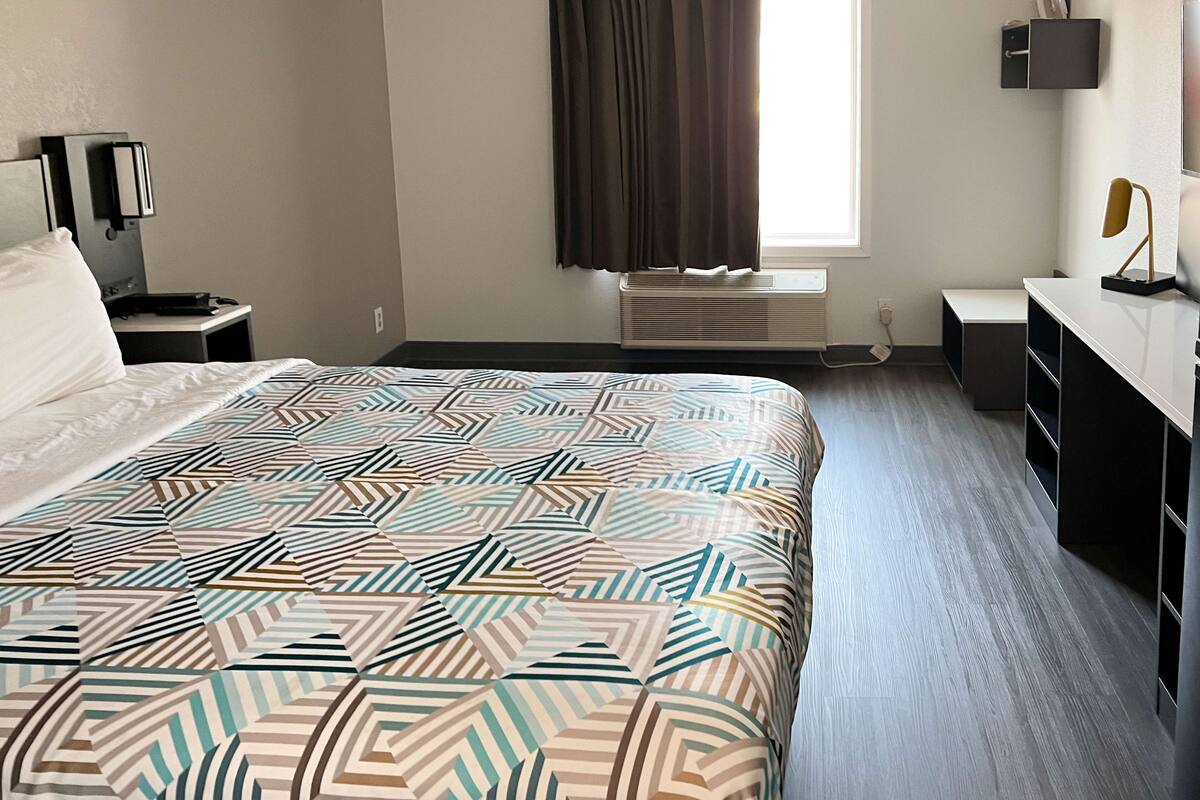 A hotel room with a large bed, a desk, a chair, a window with curtains, and a wall-mounted shelf.