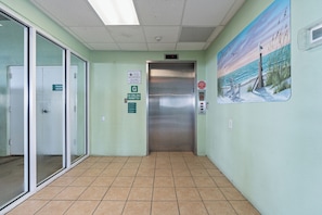 An elevator in a hallway with a beach mural on the wall, a trash room sign, and a no grilling permitted sign.