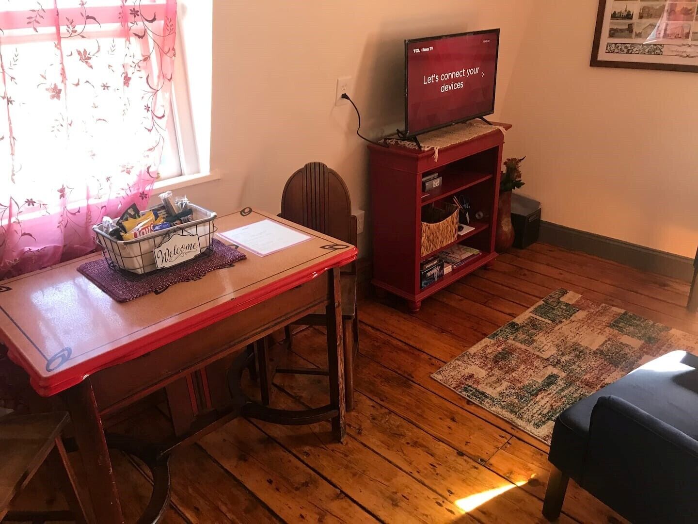 Une pièce avec un plancher en bois, une table rouge sur laquelle se trouve un panier portant l’inscription « Bienvenue », un téléviseur posé sur un meuble et un tapis.