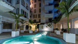 A swimming pool with tiled edges, surrounded by potted plants and a multi-story building with balconies. - Pto. Banús, modern, bright, 2-bedroom Apart., pool (Puerto Banús)