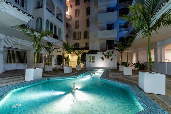 A swimming pool with tiled edges, surrounded by potted plants and a multi-story building with balconies.