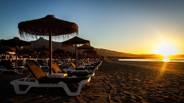 A beach with rows of sun loungers and thatched umbrellas, a clear sky, and the sun setting over the ocean. - Pto. Banús, modern, bright, 2-bedroom Apart., pool (Puerto Banús)