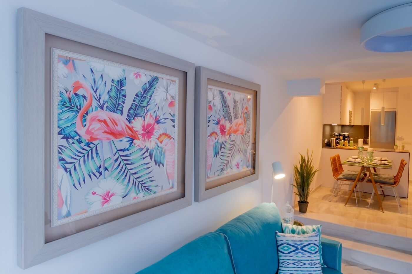 A living room with a teal sofa, a dining area with a glass table, and two framed tropical artwork on the wall.