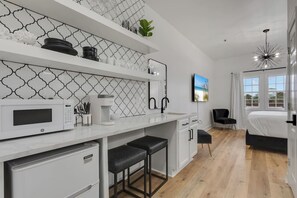 A modern kitchen with a microwave, coffee maker, and bar stools. There is a bed with a black headboard and a TV mounted on the wall.