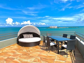 A circular outdoor seating area with a canopy, a table with chairs, and a view of the ocean and distant buildings.
