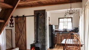 Una sala de estar acogedora con chimenea, vigas de madera en el techo, una zona de comedor con mesa y sillas, y una cocina con pared de piedra y ventana.